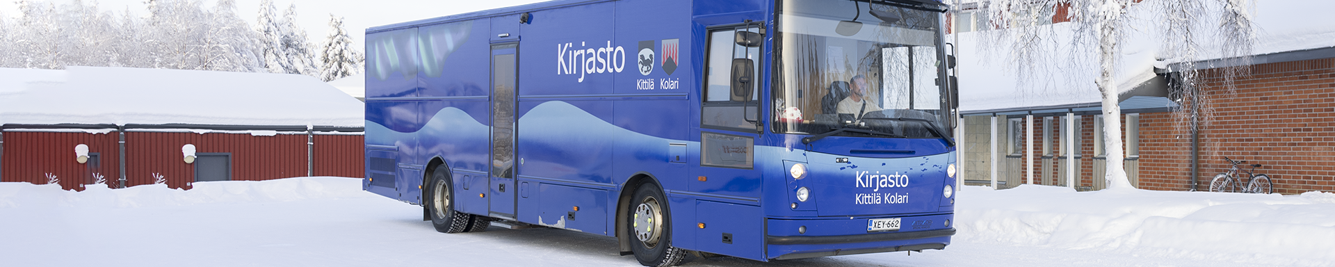 Sininen kirjastoauto seisoo lumisella pihalla talviseen aikaan. Auton kyljessä lukee “Kirjasto Kittilä Kolari”, ja kyljessä on aaltoileva vaalea kuvio. Taustalla näkyy matalia rakennuksia, joiden katot ovat paksun lumen peitossa. Puut ja maa ovat kokonaan lumen peitossa, ja yksi polkupyörä nojaa rakennuksen seinään.