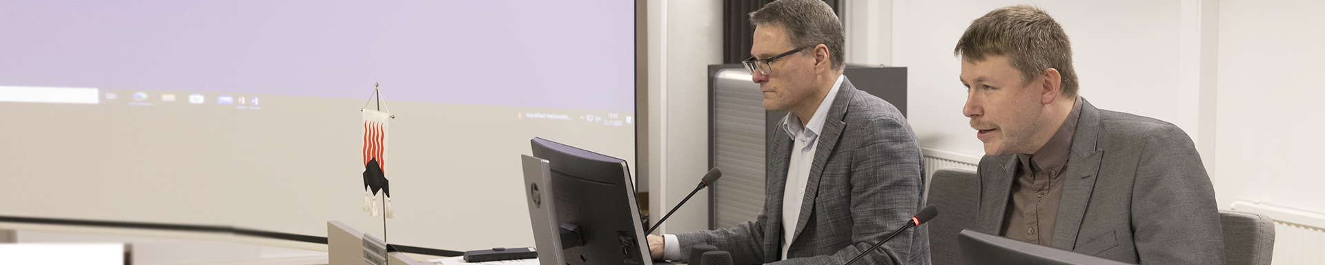 Kaksi henkilöä istuu vierekkäin neuvotteluhuoneessa pöydän ääressä, edessään tietokoneet ja mikrofonit. Taustalla näkyy suuri valkokangas, johon on heijastettu tietokoneen työpöytä. Pöydällä on myös pieni pöytälippu, jossa on punaisia ja mustia kuvioita.