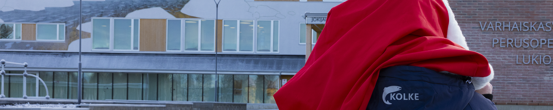 Talvinen ulkokuva koulurakennuksesta, jonka seinässä lukee "Varhaiskasvatus Perusopetus Lukio". Rakennuksen julkisivussa on suuria ikkunoita ja seinämaalauksia, joissa näkyy tunturimaisema ja eläinhahmoja. Etualalla näkyy henkilö pukeutuneena punaiseen viittaan ja tummansiniseen takkiin, jossa on valkoinen logo ja teksti "KOLKE". Taustalla luminen piha ja metallinen kaide.
