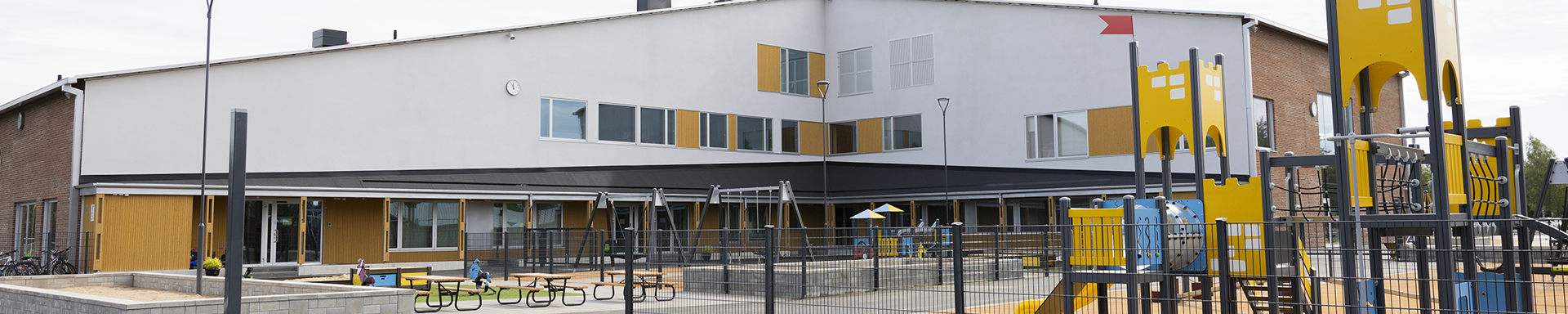 Moderni koulurakennus, jonka edustalla on leikkialue. Rakennuksessa on valkoisia ja keltaisia paneeleja sekä suuria ikkunoita, ja seinässä näkyy kello. Leikkialueella on keltaisia ja sinisiä leikkivälineitä, kuten liukumäkiä ja kiipeilytelineitä, sekä pöytiä ja penkkejä aidatulla alueella.