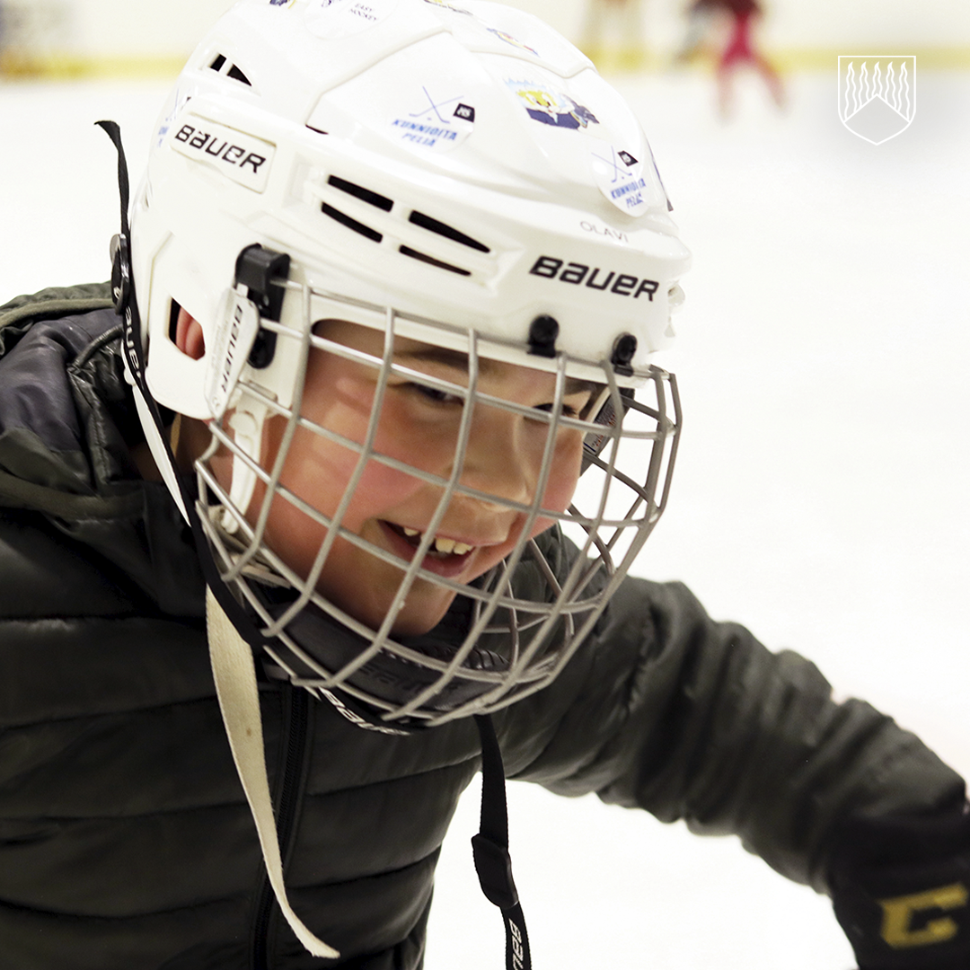 Henkilö pukeutuneena jääkiekkovarusteisiin, päässään valkoinen Bauer-merkkinen kypärä, jossa on kiinnitettynä metallinen kasvosuoja ja tarroja. Henkilöllä on tumma toppatakki ja mustat hanskat, ja hän on jääkiekkokaukalossa luistimilla. Taustalla näkyy jää ja hieman epätarkasti muita pelaajia.