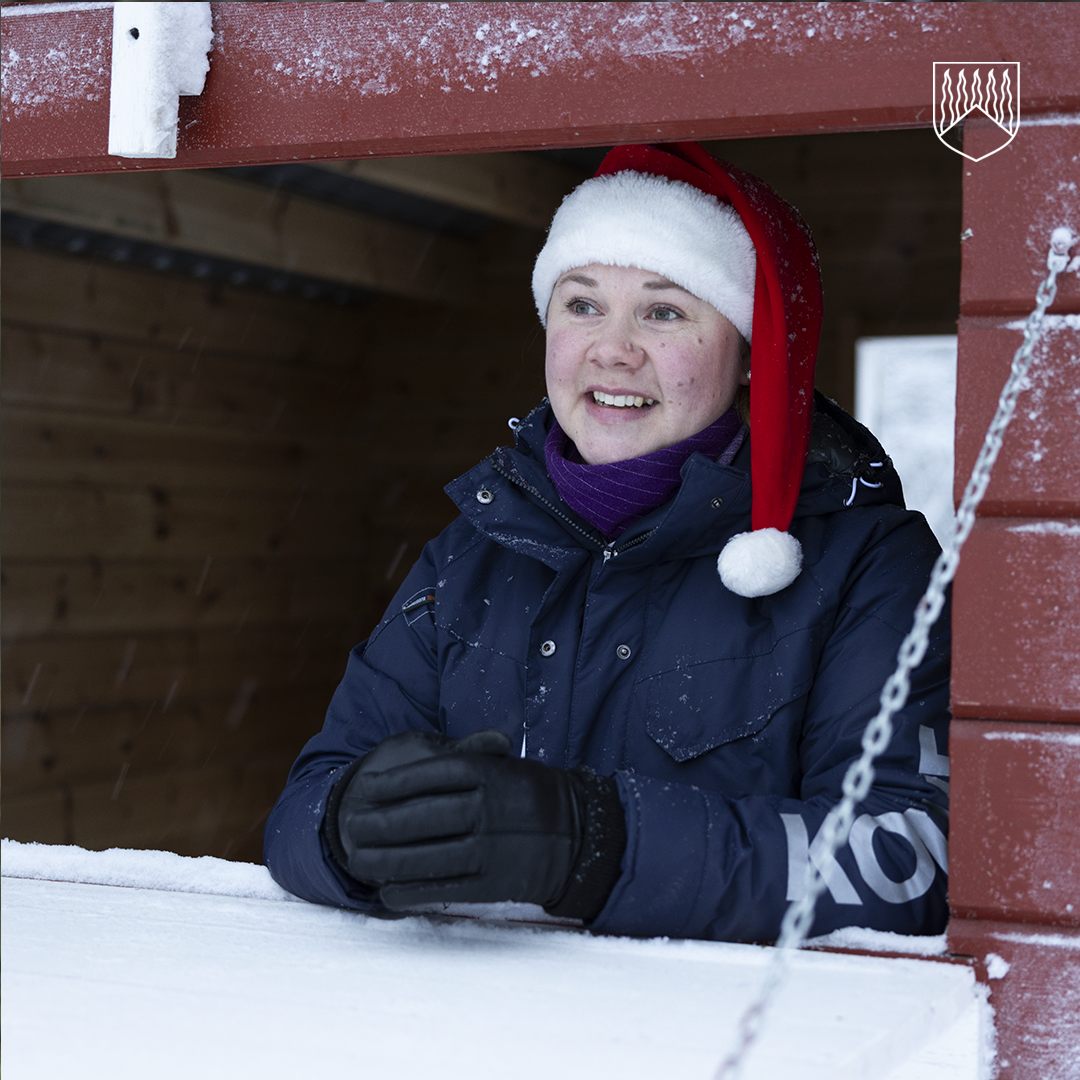 Henkilö seisoo ulkona puisessa myynti- tai palvelukojussa, pukeutuneena tummansiniseen talvitakkiin ja mustiin hansikkaisiin. Päällä on punainen joulupukin hattu valkoisella reunuksella ja tupsulla. Kojun ympärillä näkyy lunta ja jääriitteitä, ja taustalla on puinen seinä.