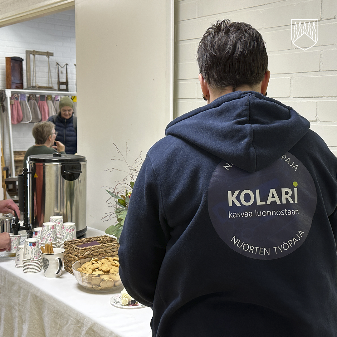 Henkilö, jolla on tummansininen huppari, jonka selässä lukee 'KOLARI kasvaa luonnostaan NUORTEN TYÖPAJA', seisoo pöydän ääressä. Pöydällä on kahvinkeitin, kertakäyttömukeja ja kulhoja, joissa on keksejä ja muita naposteltavia. Taustalla kaksi ihmistä juttelee naulakon läheisyydessä. Tila vaikuttaa olevan sisätiloissa, mahdollisesti yhteisökeskuksessa tai vastaavassa paikassa.