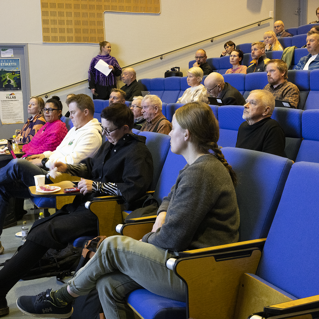 Auditoriossa istuu useita ihmisiä sinisillä kangasistuimilla, osa seuraa tilaisuutta ja osa tekee muistiinpanoja. Etualalla näkyy pöytiä, joilla on kahvikuppeja ja lautasia. Seinällä on ilmoitustaulu ja julisteita, ja tilan takaosassa seisoo henkilö pitämässä paperia kädessään.