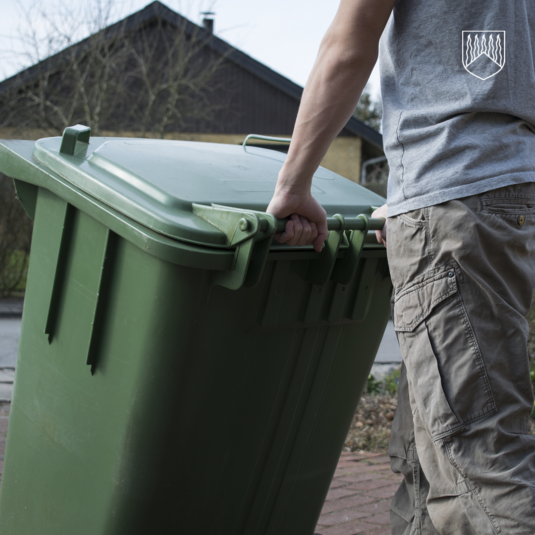 Tässä kuvassa henkilö siirtää suurta vihreää jäteastiaa pihatiellä. Henkilö tarttuu jäteastian reunassa olevaan kahvaan molemmin käsin ja kallistaa astiaa liikkeelle. Henkilöllä on yllään harmaa lyhythihainen paita, jossa näkyy pieni valkoinen tunnus, sekä vaaleat housut. Taustalla erottuu puutalo, puita ilman lehtiä sekä pihaympäristö. Valaistus on luonnonvalo, ja kuvan sävyt ovat hillittyjä.