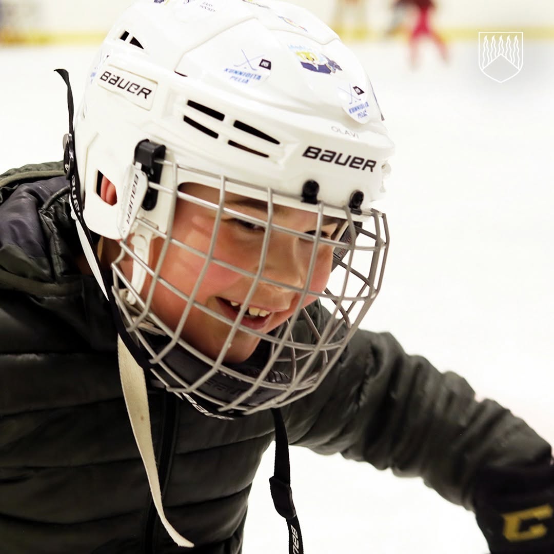 Lähikuva luistelijasta jäähallissa. Luistelijalla on valkoinen jääkiekkokypärä kasvosuojuksella ja tumma talvitakki. Taustalla näkyy jäärata ja epätarkkoja hahmoja jäällä. Kuvan oikeassa yläkulmassa on Kolarin kunnan logo.