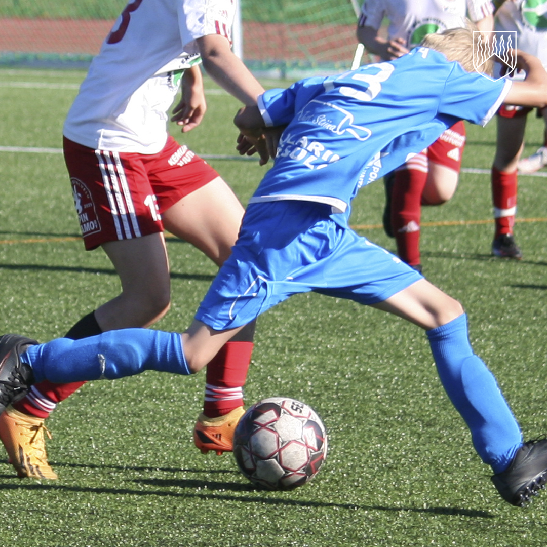 Kuvassa näkyy jalkapallo-ottelun tilanne tekonurmikentällä. Sinisiin peliasuihin pukeutunut pelaaja kurottaa kohti palloa potkaisuasennossa, samalla kun vastustajajoukkueen puna-valkoisiin asuihin pukeutuneet pelaajat pyrkivät tavoittamaan pallon. Taustalla näkyy useita pelaajia ja vihreää kenttää.