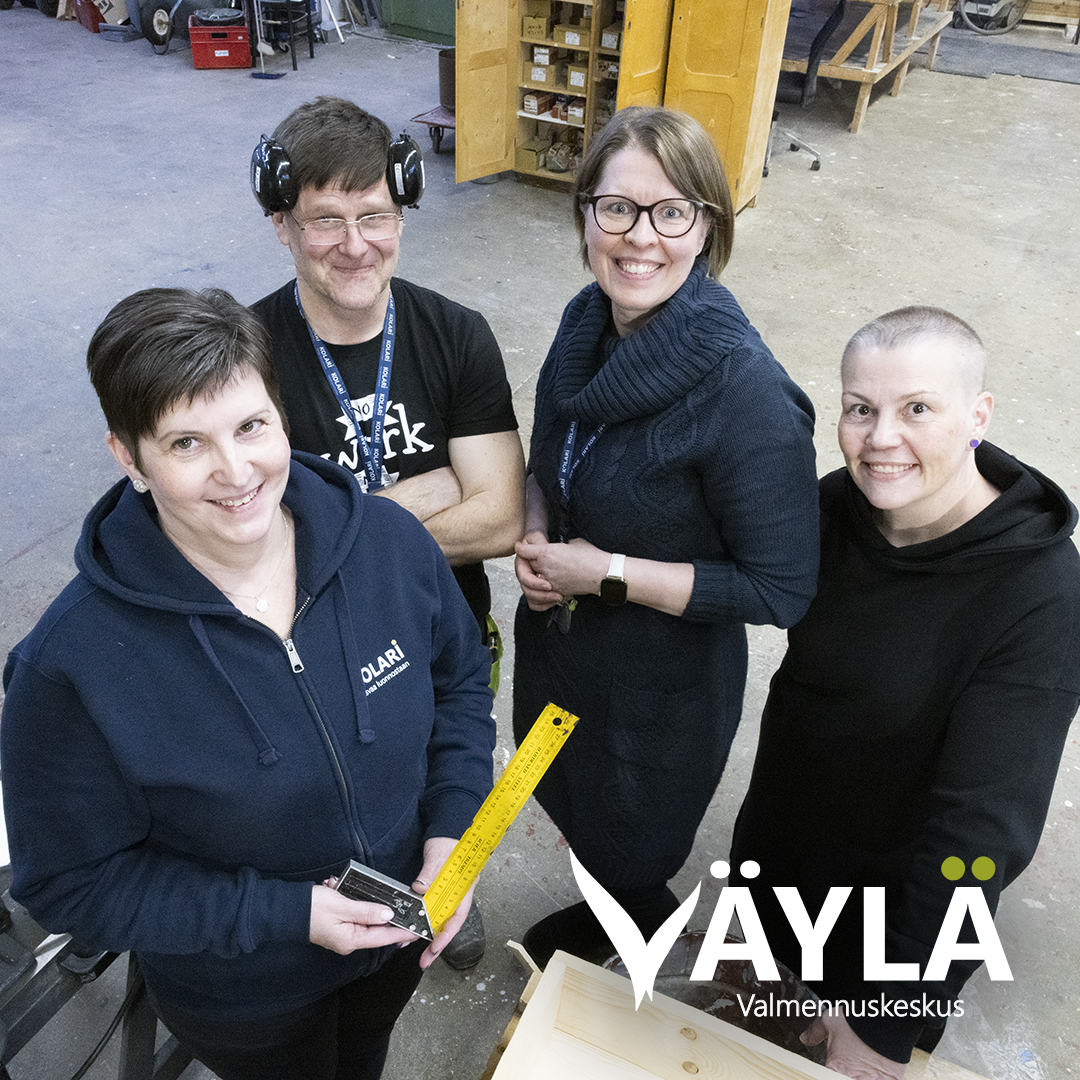 Neljä henkilöä seisoo työpajaympäristössä puutöiden parissa. Etualalla oleva henkilö pitää kädessään keltaista mittaa ja pientä työkalua. Taustalla näkyy työpöytiä, hyllyjä ja puumateriaaleja. Kuvassa on Väylä Valmennuskeskuksen logo.