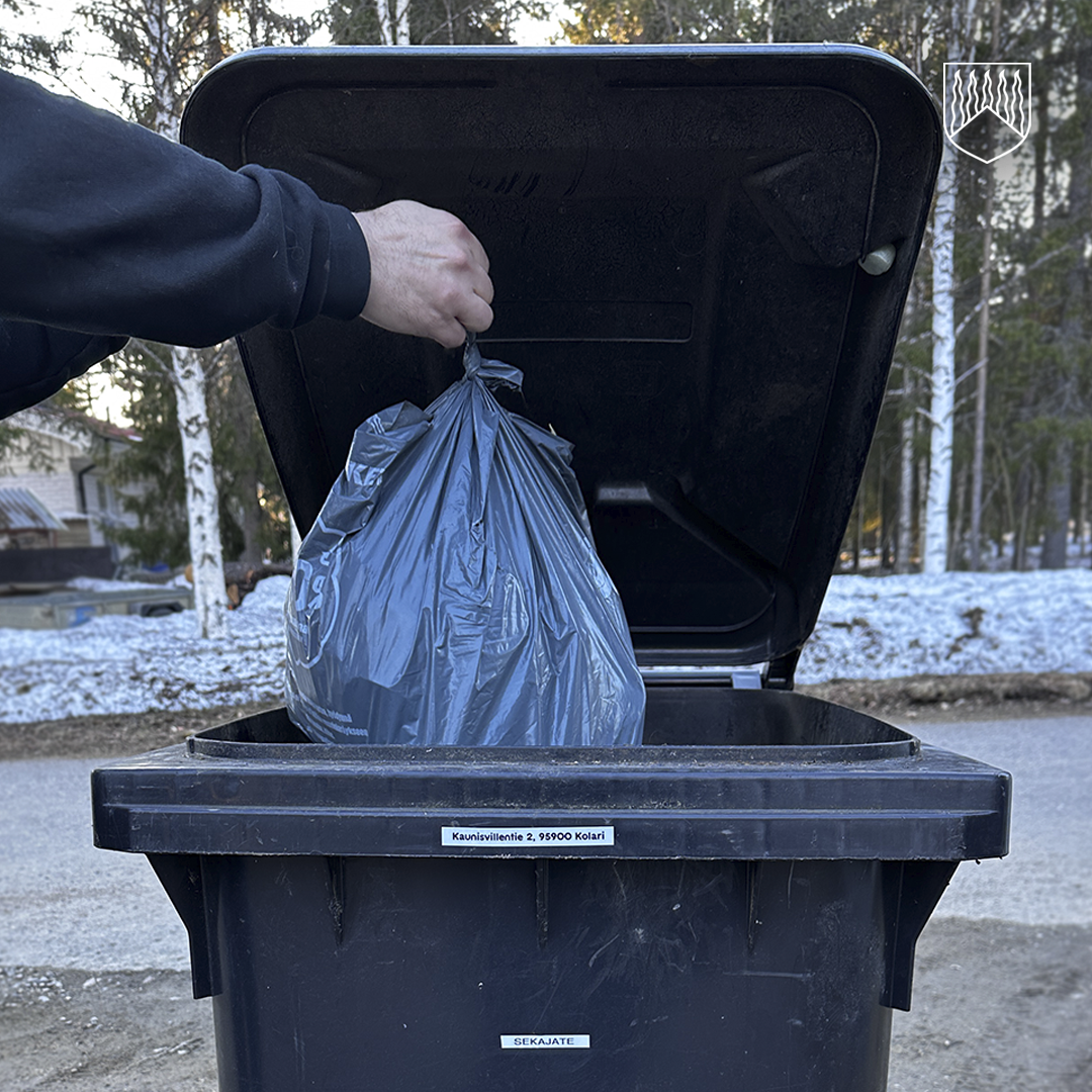 Käsi laskee sidottua mustaa jätesäkkiä avoinna olevaan mustaan jäteastiaan. Jäteastian kannessa ja etureunassa on tarrat, ja etupuolella lukee ”Sekajäte”. Taustalla näkyy asfalttipintaa sekä luminen metsäinen ympäristö.