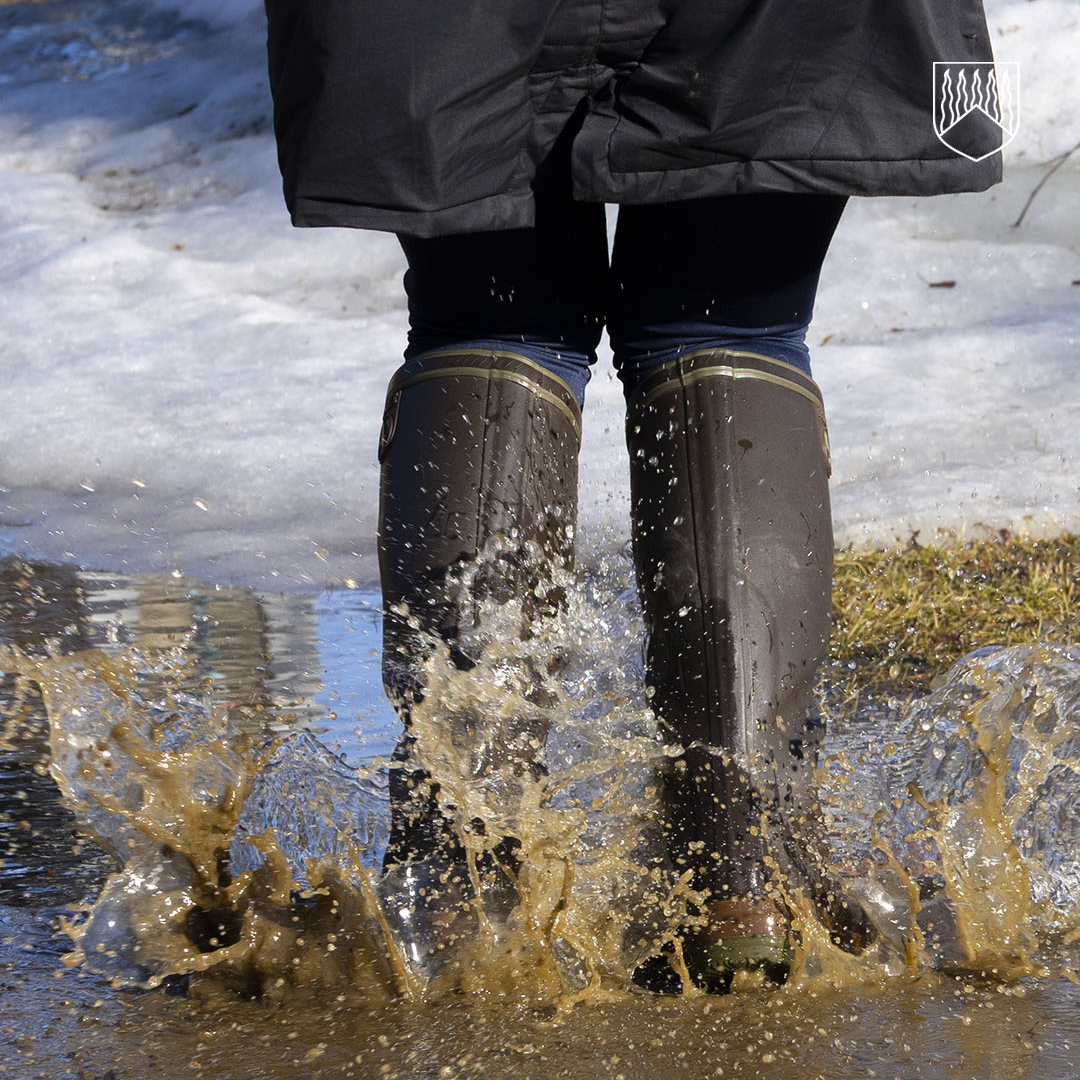 Henkilö kävelee ruskeissa kumisaappaissa sulamisvesilätäkössä, jolloin mutainen vesi roiskuu korkealle. Taustalla näkyy osittain sulanutta lunta ja keväinen nurmikon reunus.