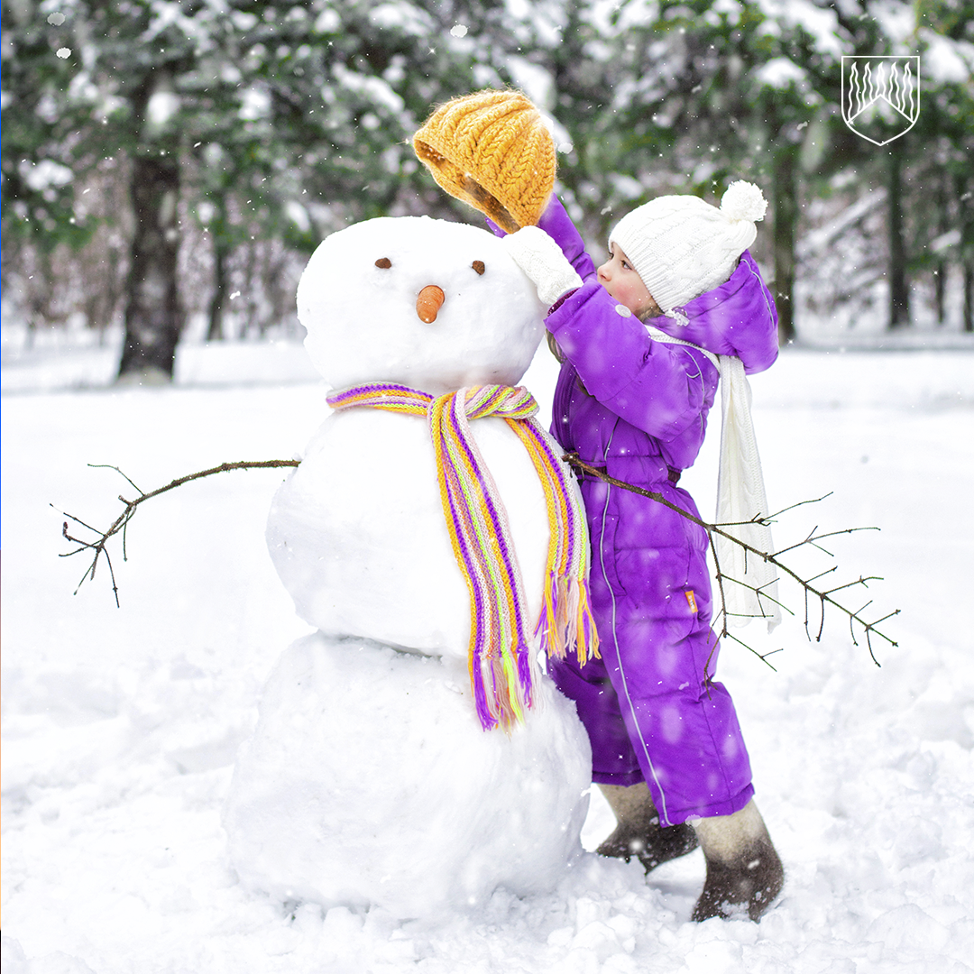 Lumiseen maisemaan rakennetun lumiukon vieressä seisoo talvivaatteisiin pukeutunut lapsi, joka asettelee lumiukolle keltaista neulepipoa. Lumiukolla on värikäs raidallinen kaulahuivi ja oksista tehdyt käsivarret. Taustalla näkyy lumisia puita.