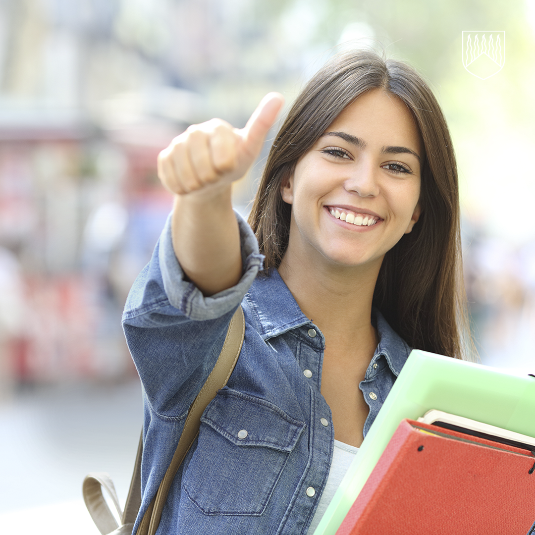 Henkilö seisoo ulkona kaupunkimaisemassa ja näyttää kädellään peukkua ylöspäin. Henkilöllä on yllään farkkukankainen paita ja olkapäällä laukku. Kädessä on useita värikkäitä kansioita. Tausta on pehmeästi sumea katuympäristö.